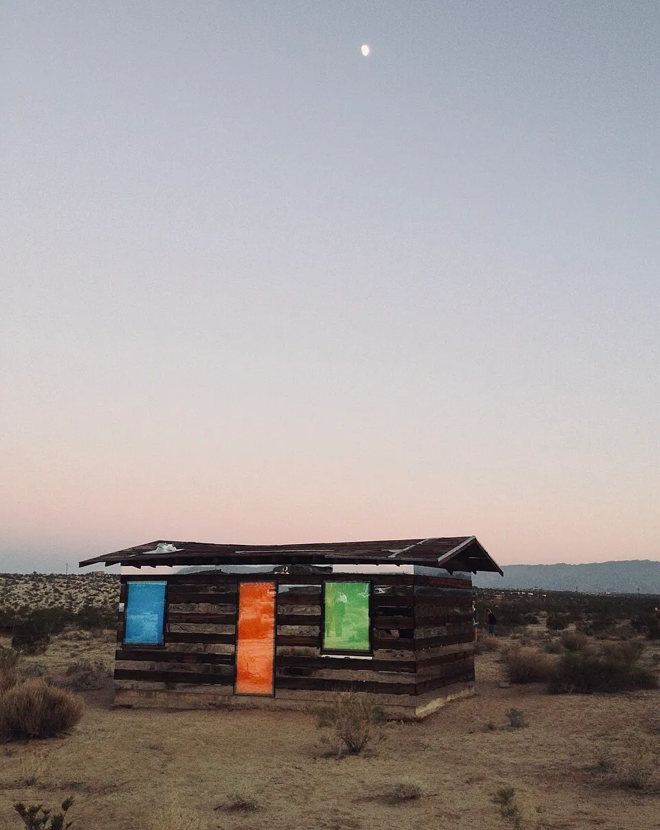 une cabane transparente en bois et en miroir lucid stead 2 Lucid Stead éclairée en bleu, orange et vert au crépuscule dans le désert