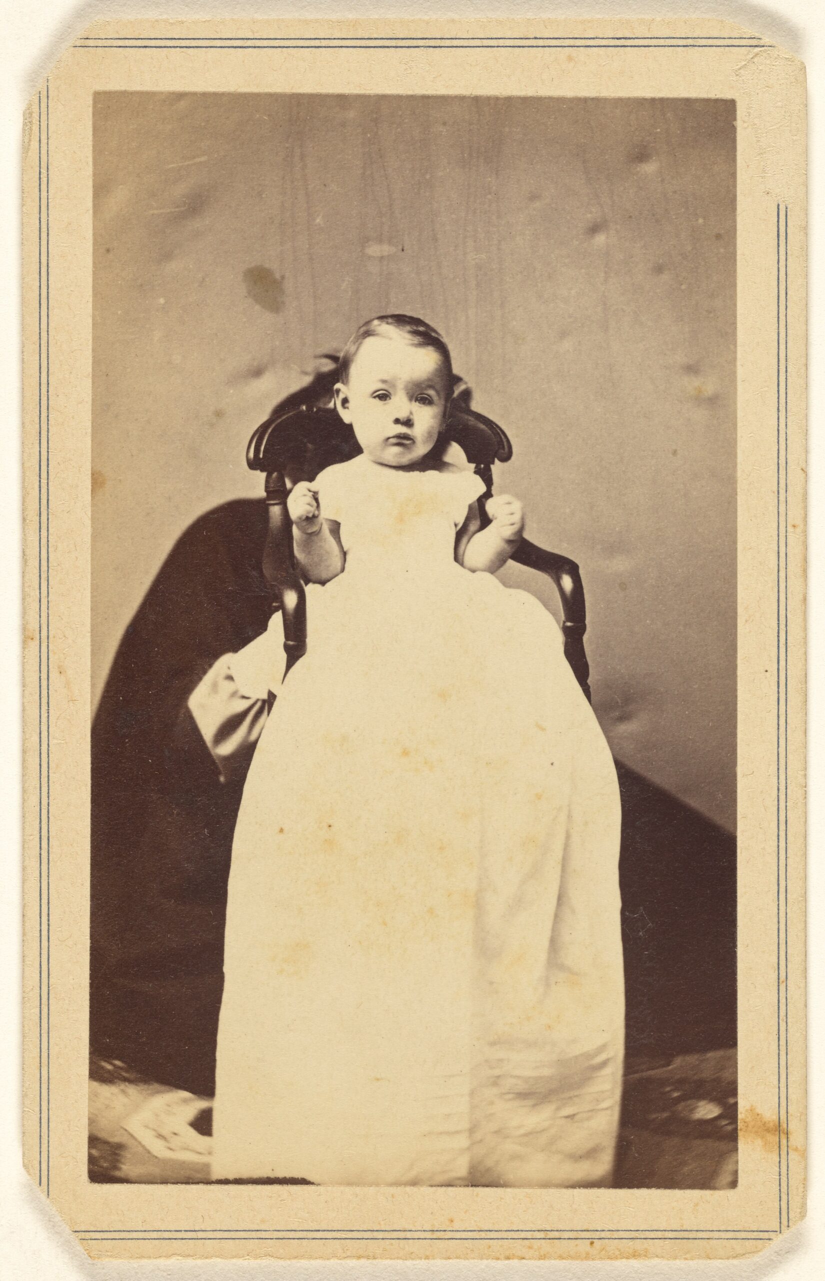 Bébé maintenu par un adulte accroupi derrière sa chaise Bébé en longue robe avec un adulte nettement visible accroupi derrière sa chaise