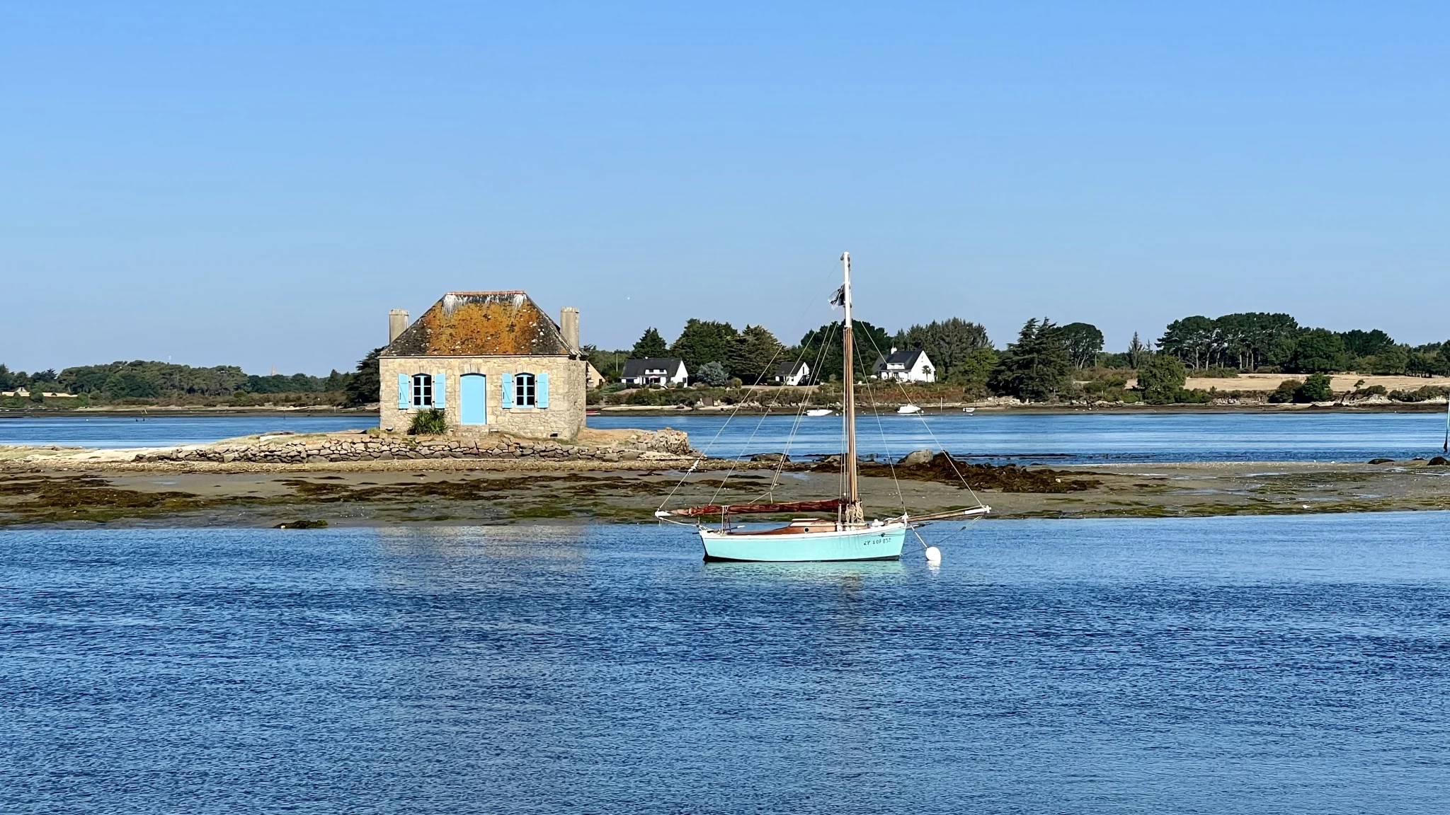 Maison de Nichtarguer aux volets bleus dans la ria d’Étel avec un voilier Maison de Nichtarguer aux volets bleus dans la ria d’Étel avec un voilier